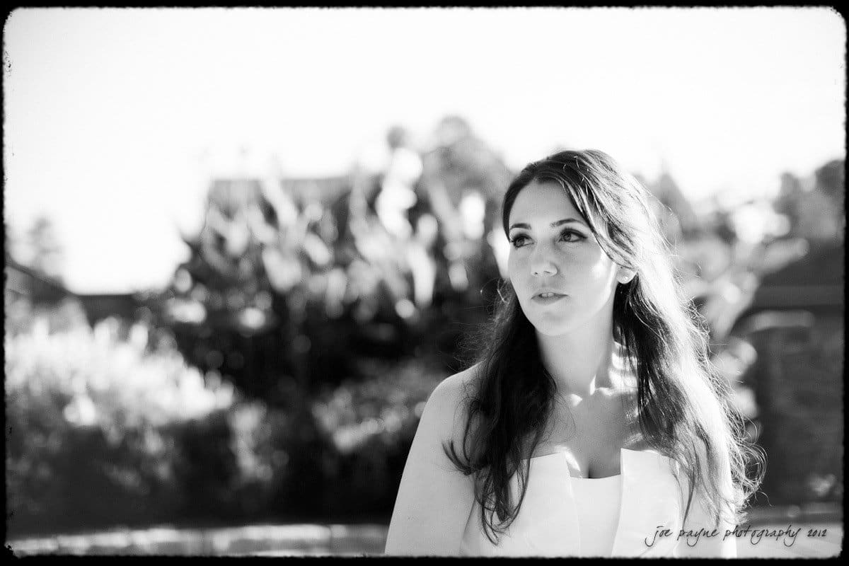 A black-and-white photo of a woman in a white dress standing outdoors. She has long, wavy hair and looks thoughtfully into the distance. The background shows blurred foliage and a clear sky.