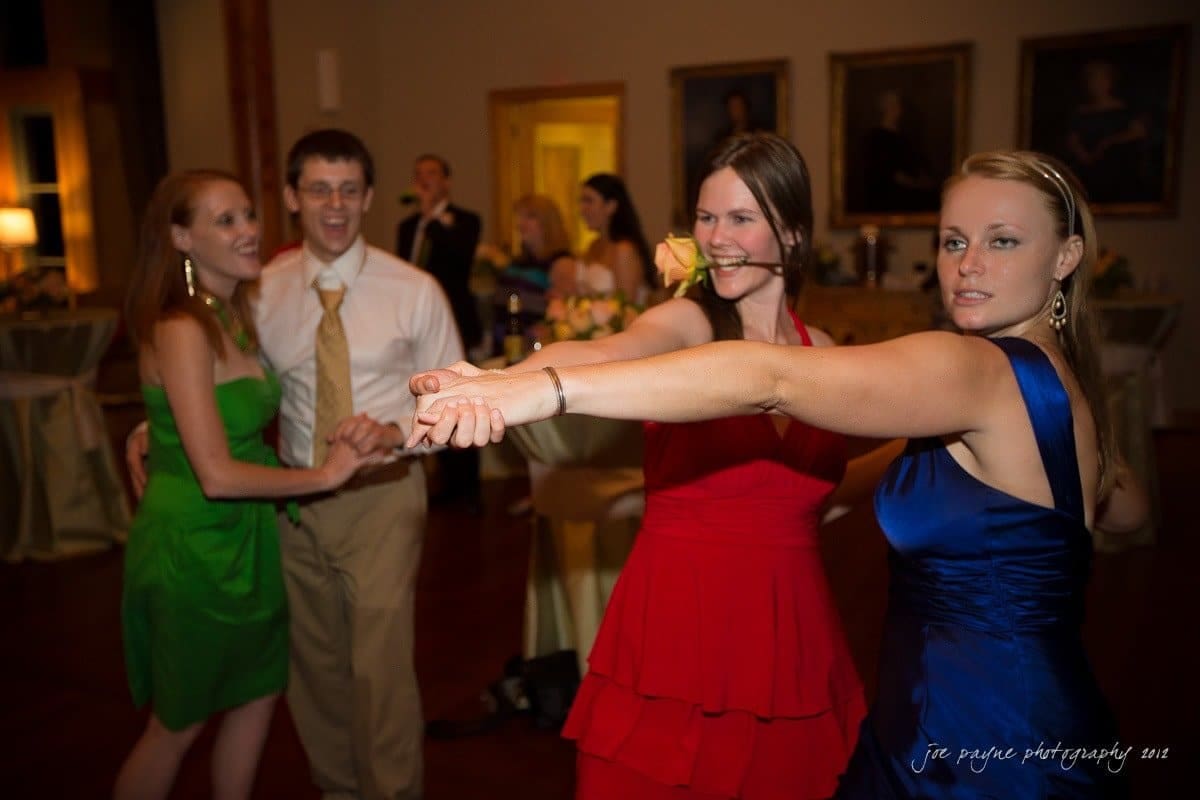 Four people are dancing and holding hands in a warmly lit room. Two women in red and blue dresses are in the foreground, while a woman in green and a man in a suit and tie are in the background, all smiling and enjoying the evening.