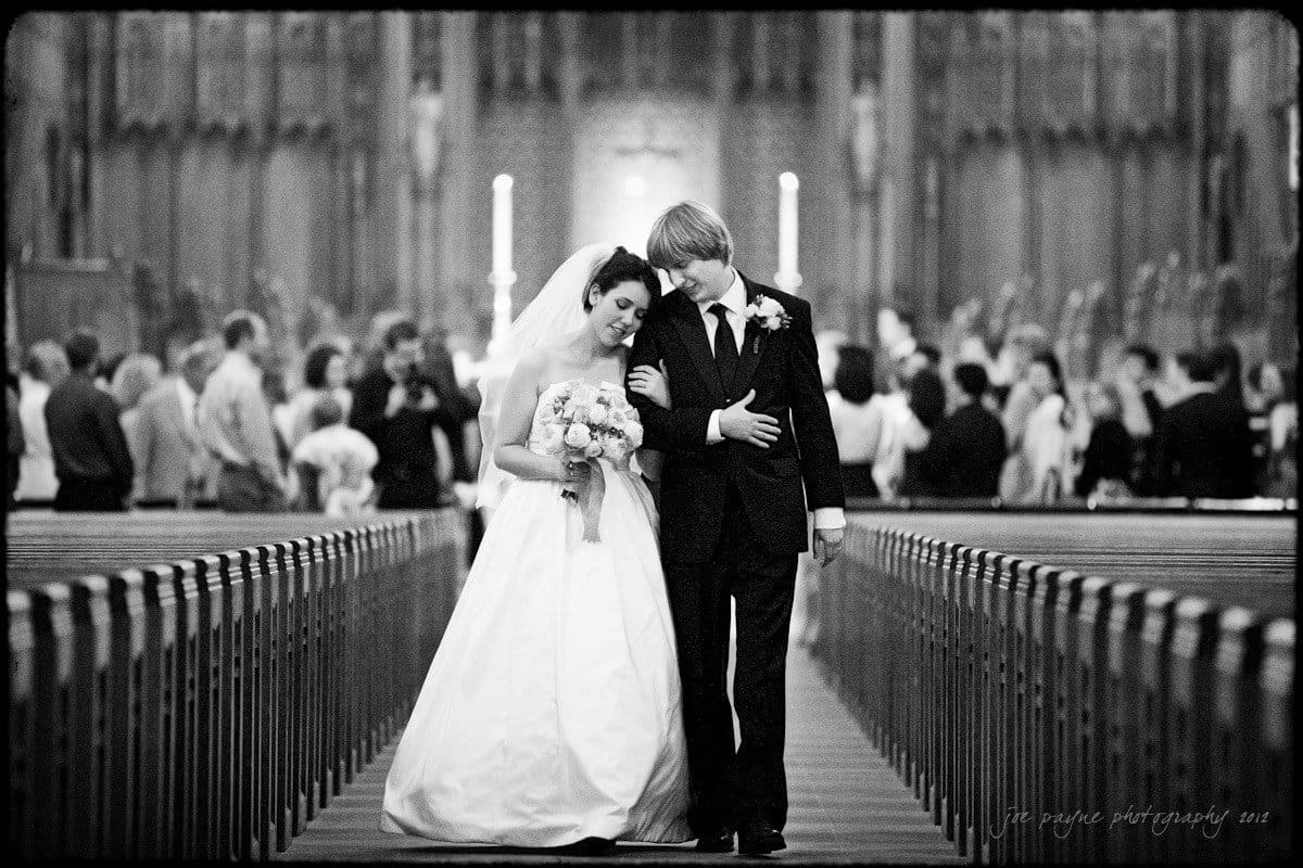 A black and white image of a bride and groom walking arm in arm down the aisle of a church. The bride is wearing a long white gown and holding a bouquet. The groom is in a suit. The background shows blurred guests seated.