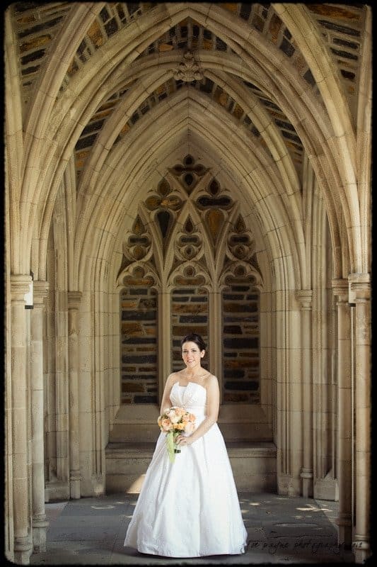 A bride in a white gown holds a bouquet while standing under a stone archway with intricate gothic architecture. The scene conveys elegance and serenity.
