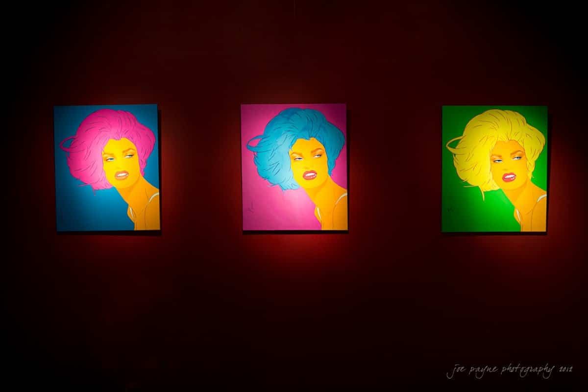 Three colorful pop art portraits of a person with vibrant hair in magenta, blue, and yellow are displayed on a dark wall. Each portrait is lit from above, highlighting the bold colors and expressive features.