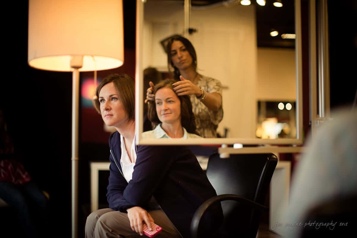 A woman sits in a salon chair holding a phone, looking at her reflection. Another woman stands behind her, styling her hair. A large mirror and soft lighting create a warm ambiance.