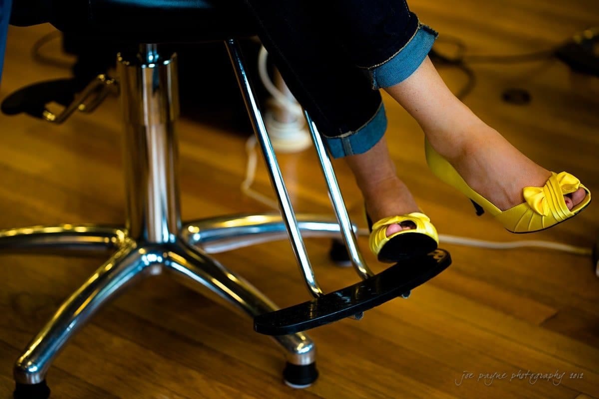 A person wearing yellow high heels sits on a salon chair with metal footrests. The floor is wooden, and the person is dressed in dark jeans.