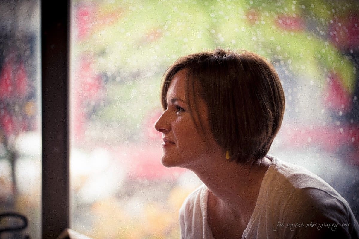 A person with short hair gazes out of a window on a rainy day. The blurred background shows raindrops on the glass and a mix of green and red hues outside. The warm indoor lighting highlights the profile of their face.