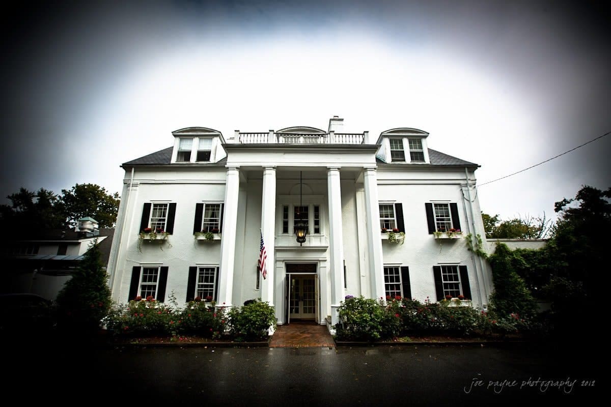 A large white mansion with tall columns and dormer windows. An American flag hangs near the entrance. The garden is lush, with colorful flowers bordering a pathway leading to the door. The sky is overcast.