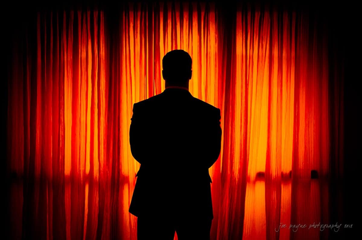Silhouette of a man in a suit standing before bright orange curtains, creating a dramatic contrast. The lighting emphasizes his outline against the glowing curtain backdrop.
