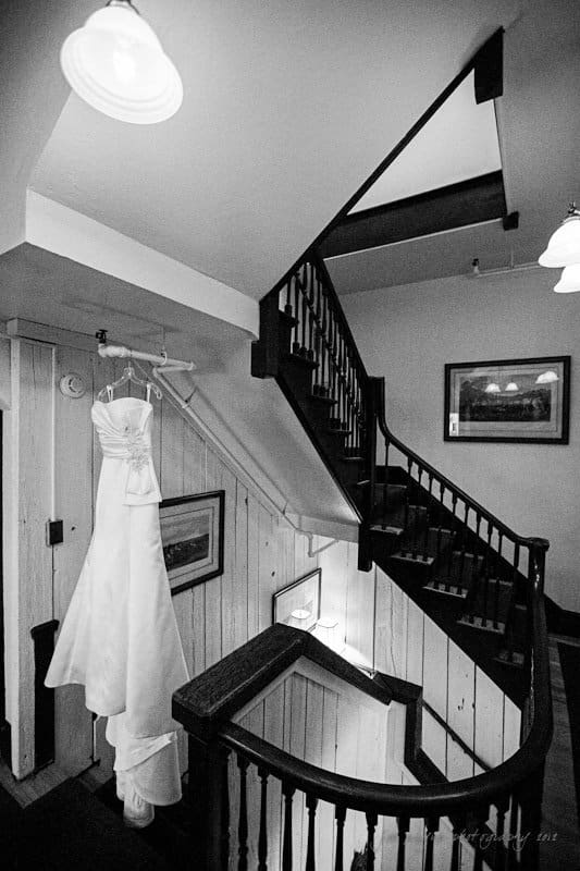 A black and white photo of an elegant wedding dress hanging on a wooden hanger by a rustic staircase. The wooden railing and framed artwork on the wall add a cozy, vintage charm to the setting.