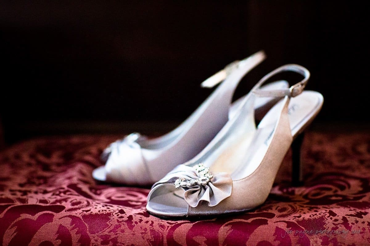 Elegant satin high-heeled shoes with open toes and decorative bows rest on a rich, patterned burgundy fabric. The shoes feature delicate embellishments, creating a sophisticated and classy appearance.