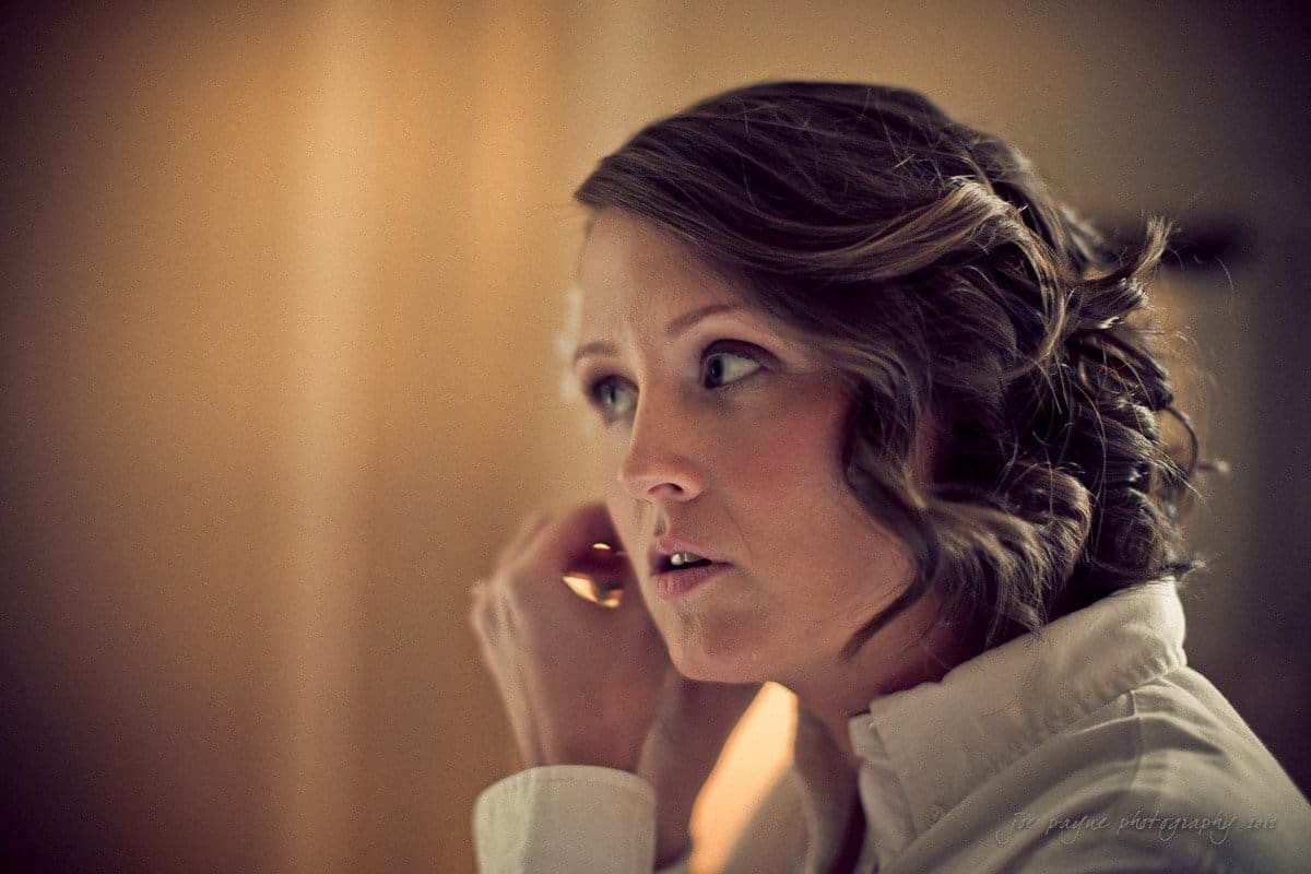 A woman with styled hair is adjusting an earring. She is wearing a white shirt and has a focused expression. The background is softly blurred.