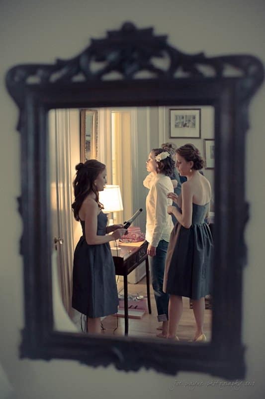 A mirror reflection shows three women preparing for an event. One is styling her hair with a curling iron, another is holding a drink, and the third wears a floral headpiece. They are dressed in formal attire, standing in a softly lit room.