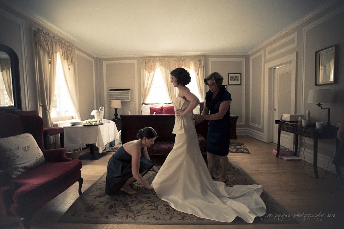 A bride gets ready in a vintage-style room with two women assisting her. One woman adjusts the train of her gown while the other helps with the dress. Sunlight streams through the windows, illuminating the elegant setting.