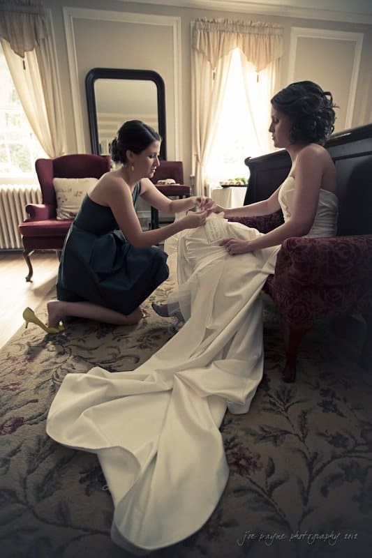 A woman in a dark dress kneels and adjusts the shoe of a seated bride wearing a white gown with a long train. They are in an elegant room with vintage furniture and a large mirror.