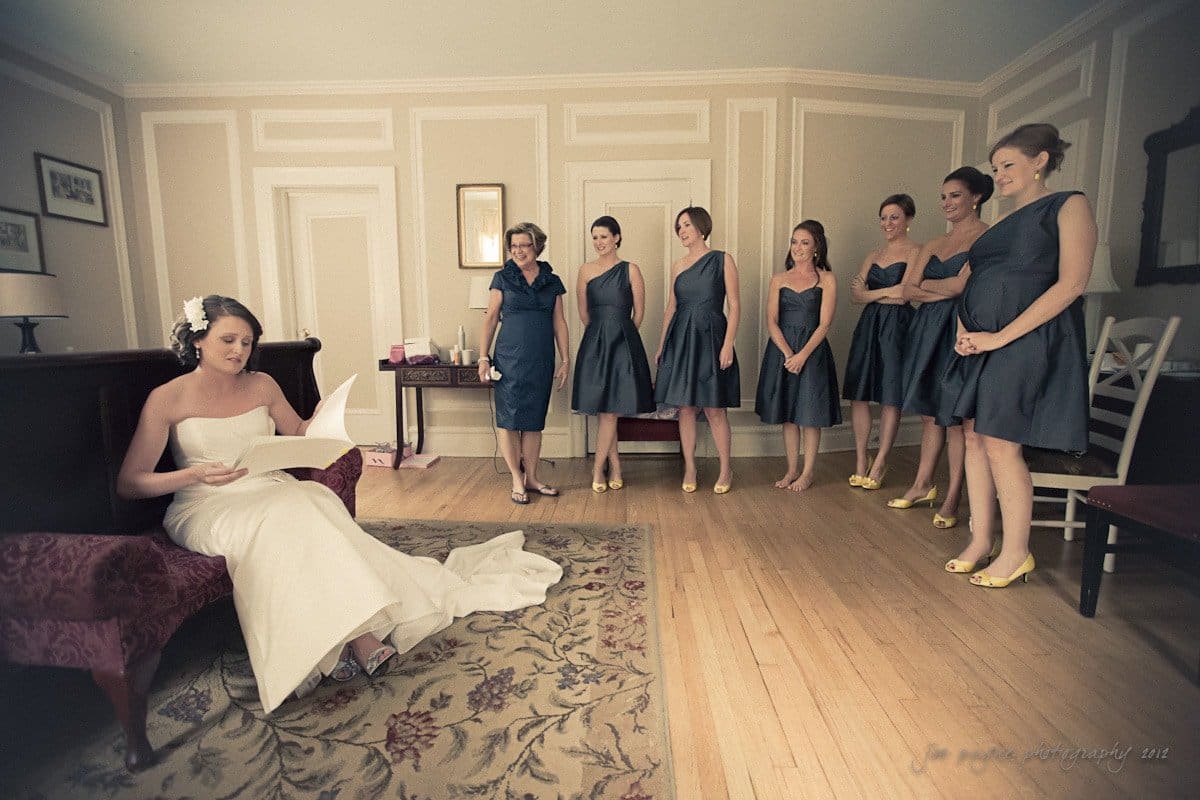 A bride in a white dress sits and reads a letter in a room with wooden floors. Six bridesmaids in matching teal dresses and yellow shoes stand nearby, observing. The setting appears to be a formal room with light-colored walls and framed art.