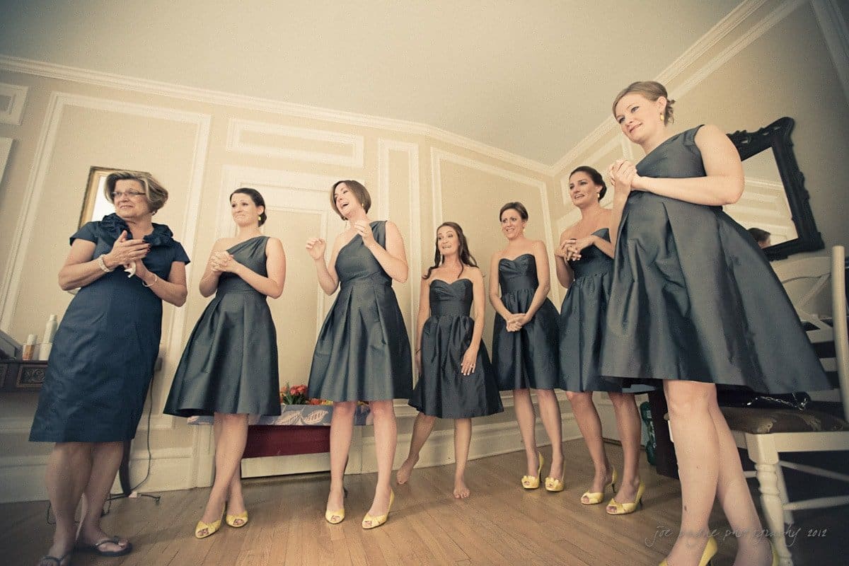 A group of seven women in matching dark dresses stands in a well-lit room with beige walls and wooden flooring. They appear to be preparing for a formal event, possibly a wedding. A mirror and a chair are visible in the background.