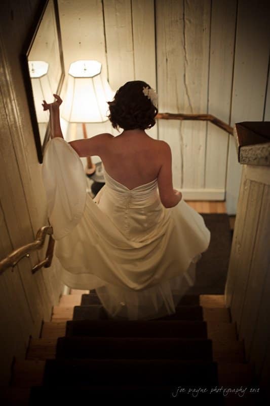 A woman in a white wedding dress descends a dimly lit wooden staircase. She holds up her dress with one hand, and a soft light from a lamp illuminates the scene. The walls are textured with a rustic finish.