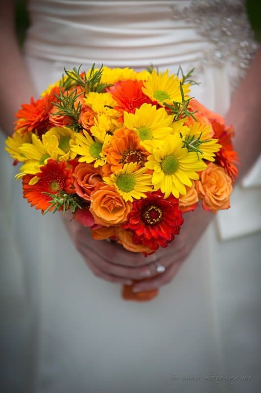 A close-up of a bride holding a vibrant bouquet of yellow, orange, and red flowers, including daisies and roses. The flowers contrast vividly against her white wedding dress.