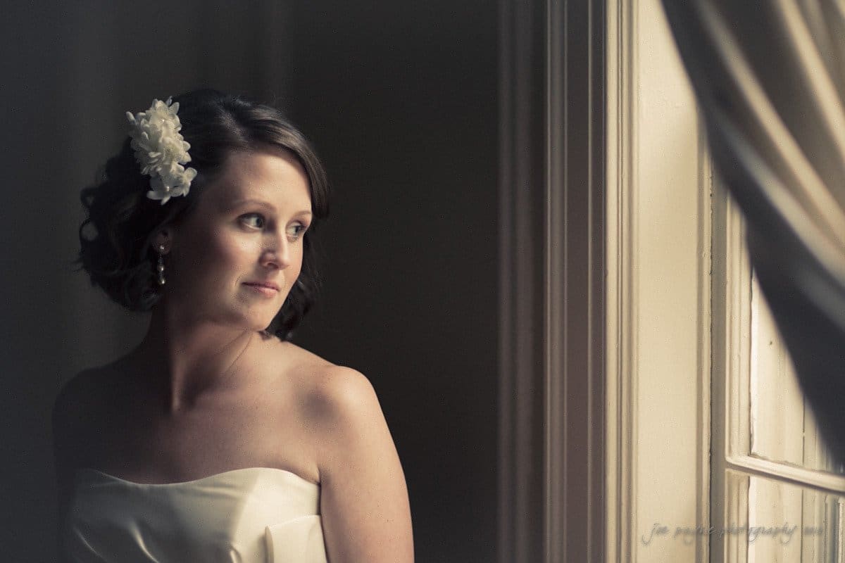 A bride in a strapless white gown stands by a window, gazing contemplatively outside. She has a white flower in her short hair and soft lighting highlights her peaceful expression against the neutral background.