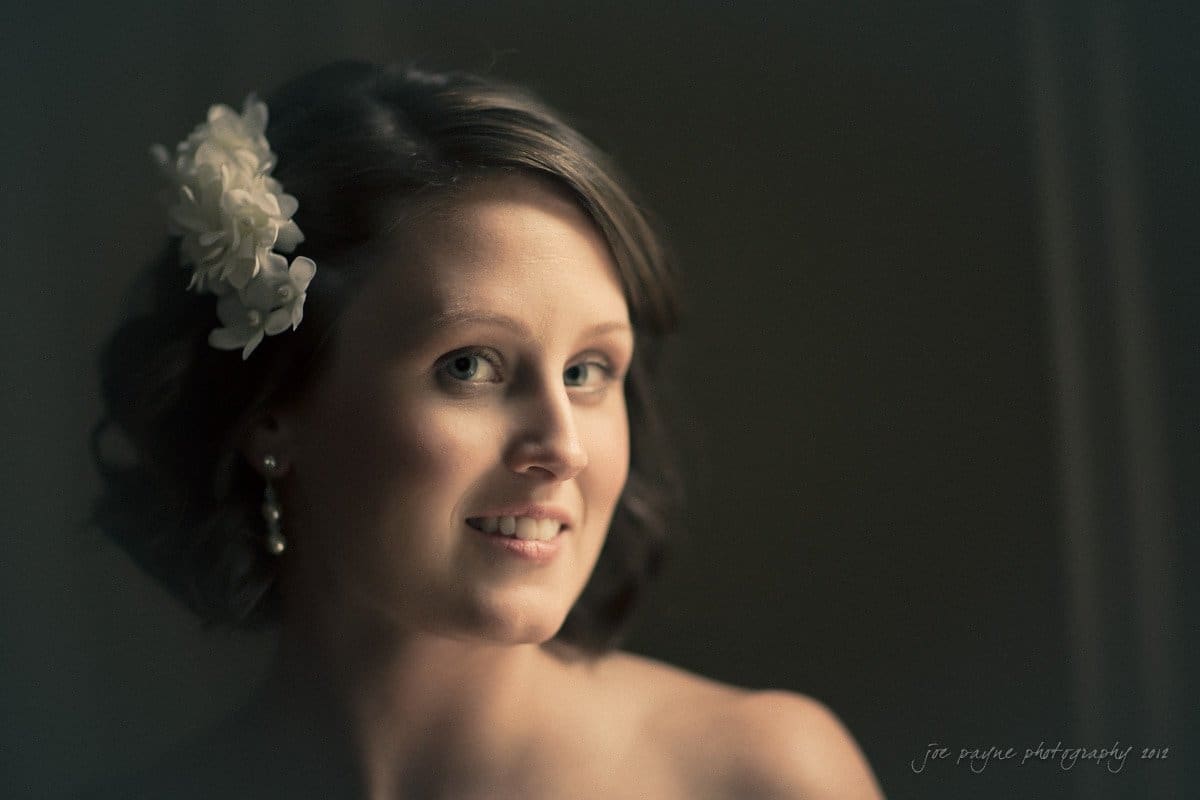 A woman with short brown hair smiles softly, wearing a delicate white flower in her hair. The background is softly blurred, highlighting her elegant earrings and subtle makeup in gentle lighting.
