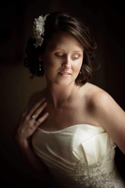 A bride in a white strapless dress and floral hair accessory gazes downward, softly smiling. The lighting creates a serene and intimate atmosphere, highlighting her elegant features and gentle pose.