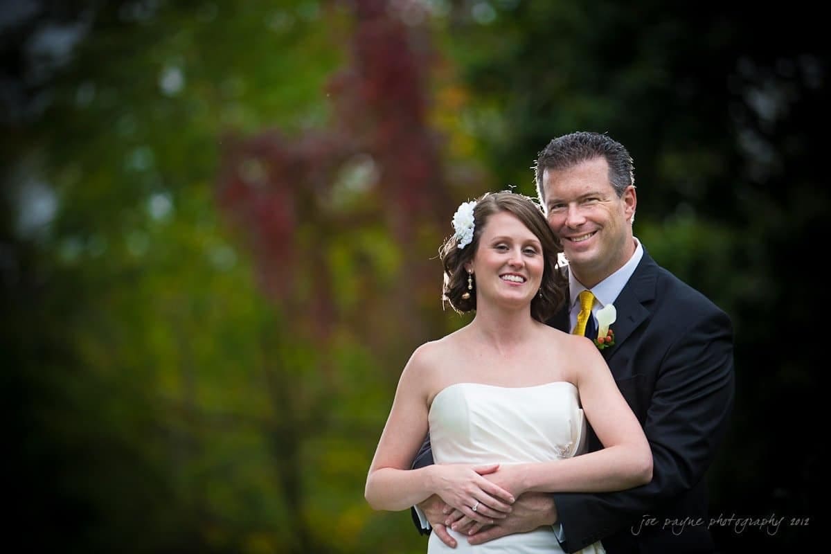 A bride and groom are standing outdoors, smiling and embracing. The bride is wearing a white strapless dress and a floral hairpiece, while the groom is in a dark suit with a boutonniere. The background is a blurred garden setting.