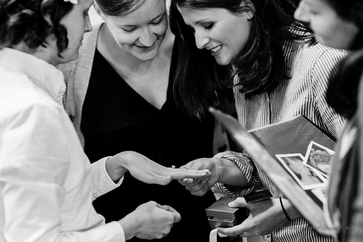A black and white photo shows a group of women gathered around, smiling and examining a ring on one persons hand. One woman holds a small box, possibly for the ring. Their expressions show joy and excitement.
