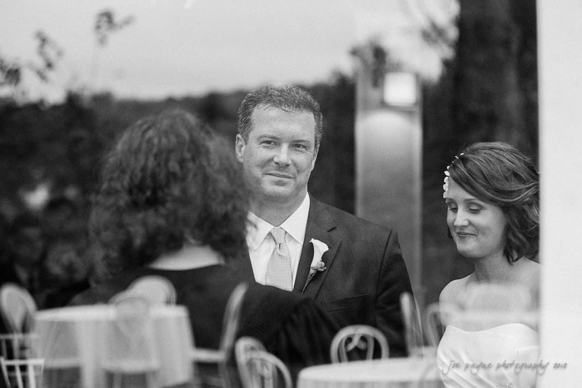 A black-and-white photo shows a bride and groom during a wedding ceremony, captured through a window. The groom wears a suit and boutonniere, while the bride wears a strapless dress with a hair accessory. Their expressions are solemn and reflective.
