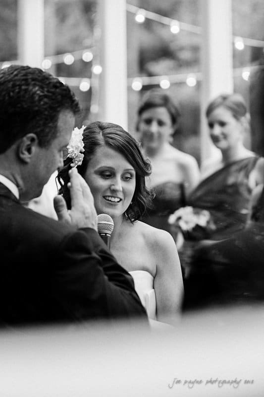 A black and white photo of a wedding ceremony. A bride is holding a microphone, smiling, and facing a man who appears to be speaking. Bridesmaids can be seen in the background. Strings of lights hang above, creating a festive atmosphere.