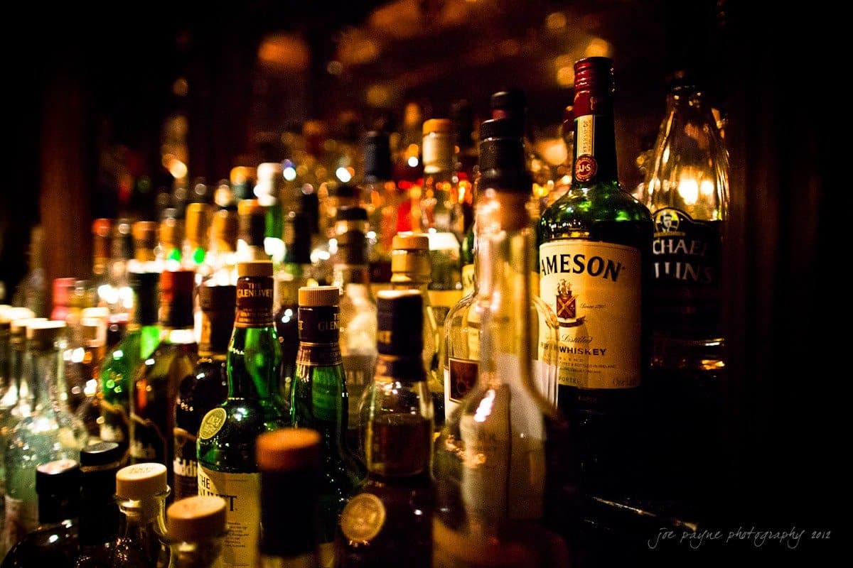 A dimly lit bar shelf is filled with a variety of liquor bottles. The labels are visible, featuring whiskey, rum, and other spirits. The warm lighting creates a cozy and inviting atmosphere.