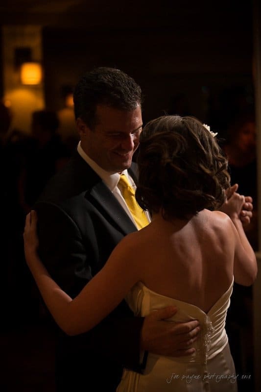 A couple shares a romantic dance in dim lighting. The man wears a suit with a yellow tie, and the woman is in a strapless white dress. They hold each other closely, surrounded by a warm, intimate atmosphere with soft-focus background lights.