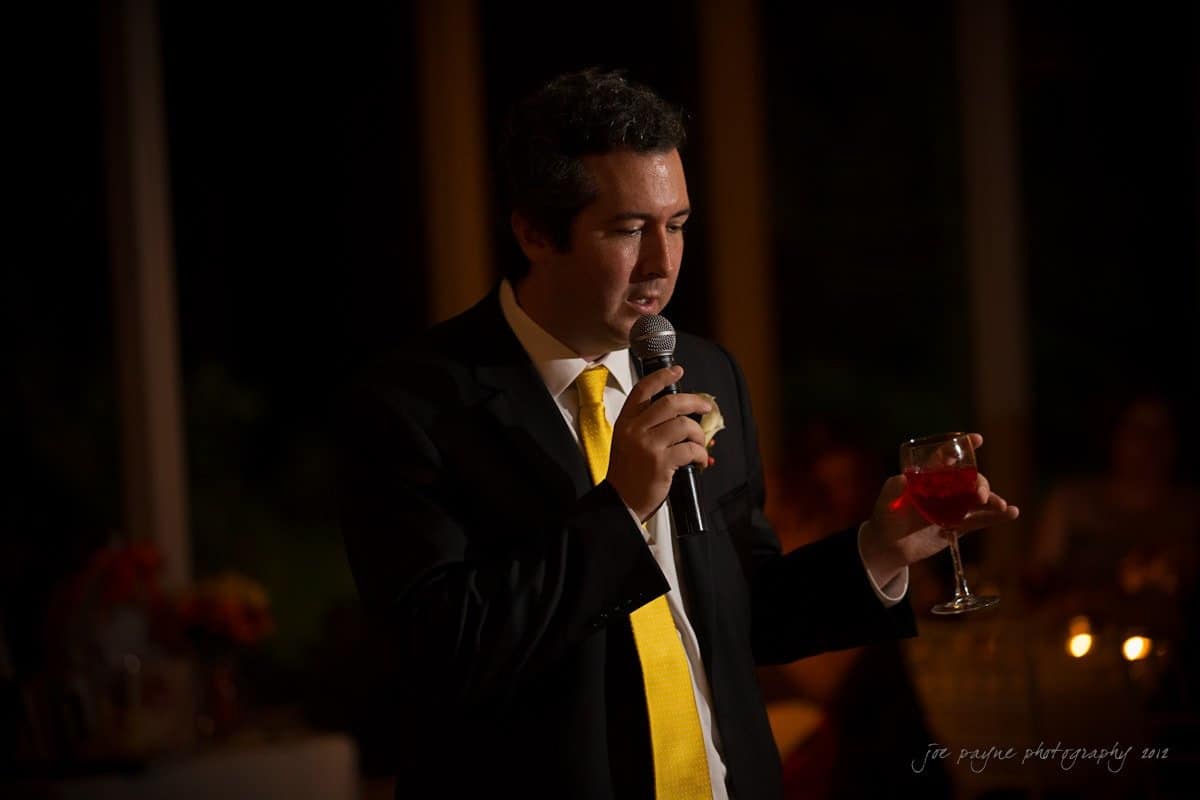 A man in a suit and yellow tie holds a microphone in one hand and a glass of red liquid in the other. He appears to be speaking at an indoor event. The background is dimly lit with blurred figures seated at tables.