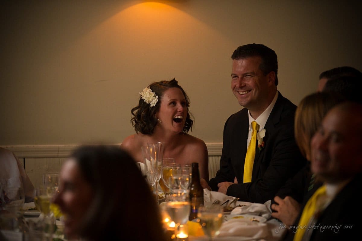 A woman in a wedding dress and a man in a suit with a yellow tie are sitting at a table, laughing and enjoying a wedding reception. They are surrounded by other guests in dim lighting with glasses and plates on the table.
