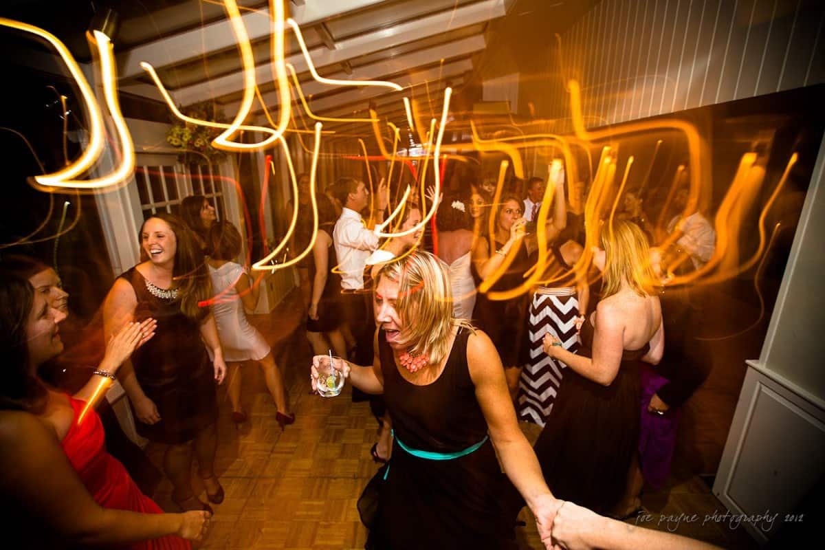 A lively party scene with people dancing energetically. Blurred lights create a dynamic, vibrant atmosphere. Attendees in formal attire are enjoying themselves on the dance floor, with one person holding a drink.