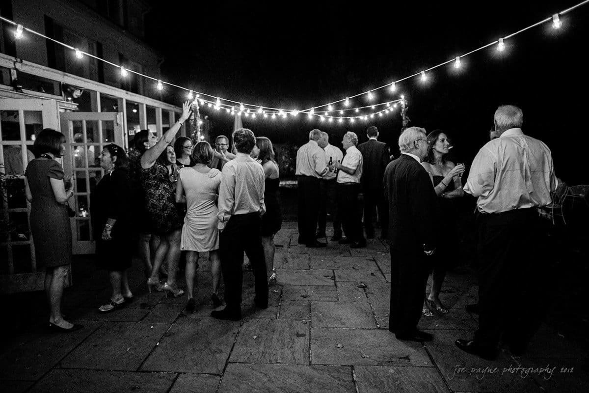 A black and white photo of a nighttime outdoor gathering. People are talking and mingling under string lights on a patio. Some are holding drinks, and others appear to be dancing or conversing in groups.