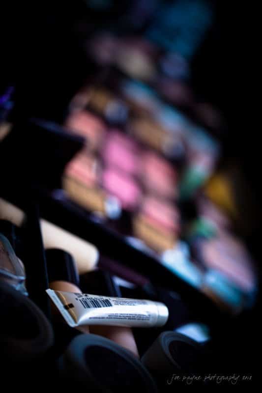 A close-up of various makeup items, including tubes and an assortment of brightly colored eyeshadow palettes. The image is artistically blurred with a focus on the foreground items.