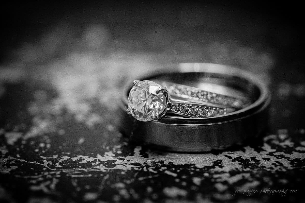 Close-up of an engagement ring with a large central diamond set in a band with smaller diamonds, resting on a wedding band. Both rings are placed on a textured surface, captured in black and white.