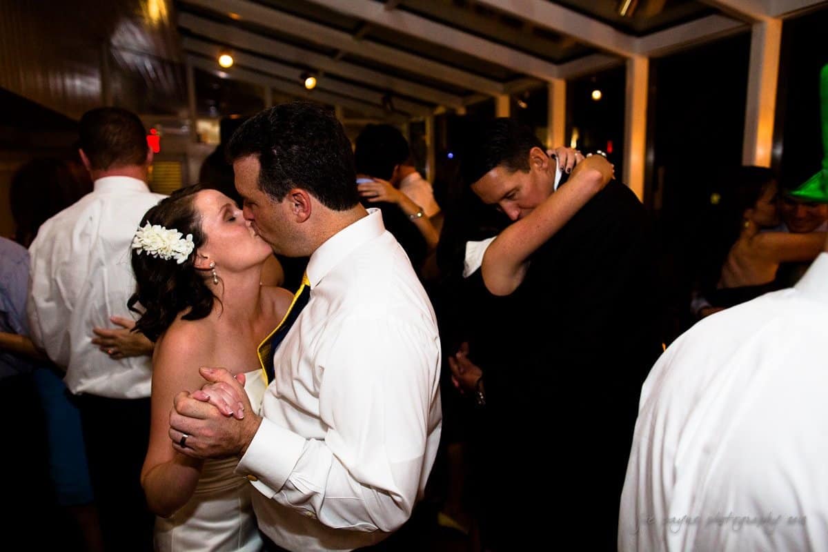A couple in wedding attire dances closely, sharing a kiss. Other couples are also dancing in the background. The room is softly lit, creating a romantic atmosphere.