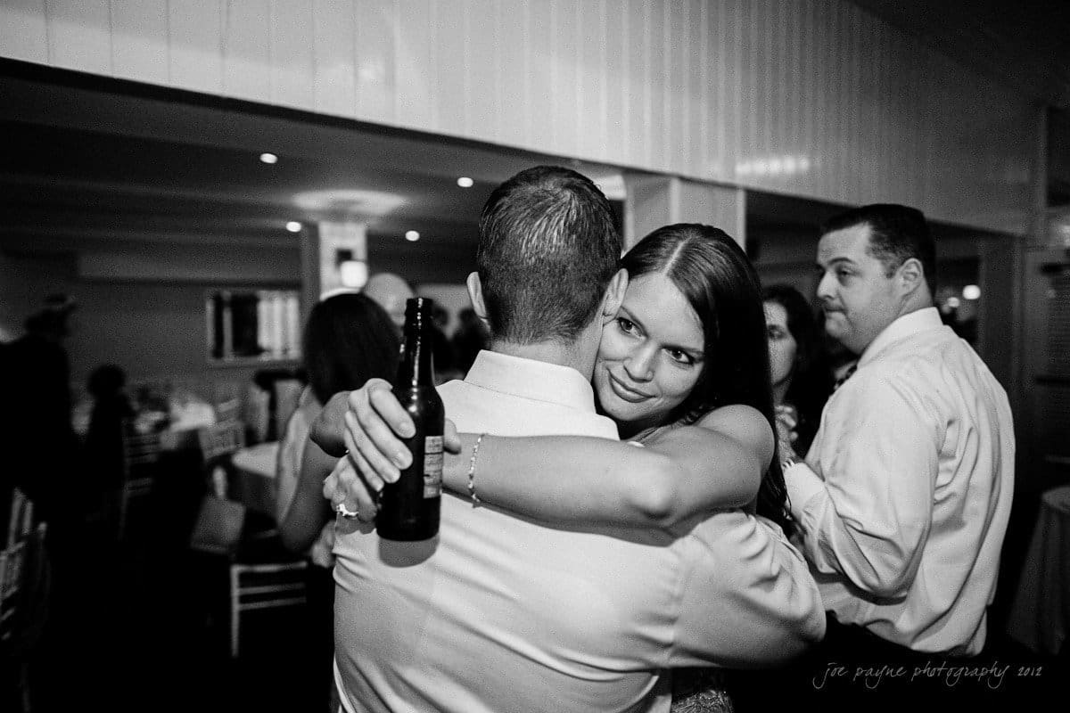 A woman embracing a man indoors, holding him closely with her eyes open. The mans back is facing the camera. Another man stands nearby holding a drink. The setting appears to be a social gathering or event.