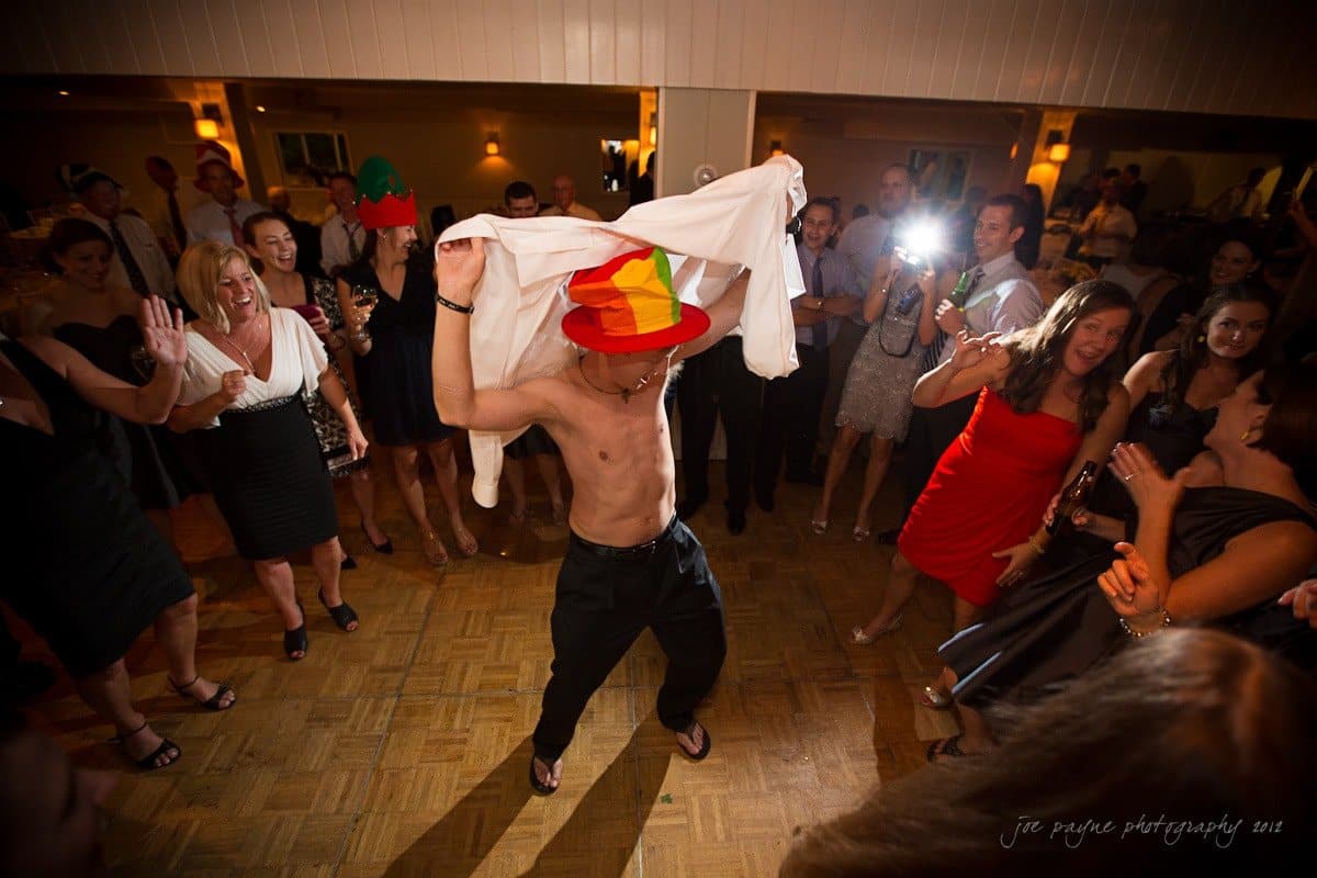 A shirtless person wears a colorful hat while dancing energetically in the center of a crowd at a party. Onlookers are clapping and smiling, some holding drinks. The room is dimly lit, creating a lively and festive atmosphere.