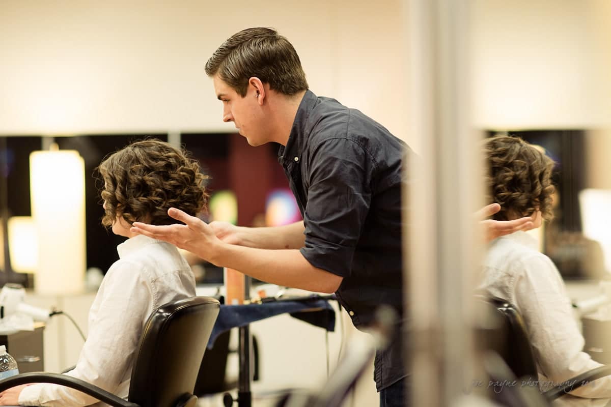 A hairstylist is working on a seated clients hair in a salon. The client has short, curly hair, and the stylist is gesturing with his hands. The background shows blurred salon lighting and mirrors.