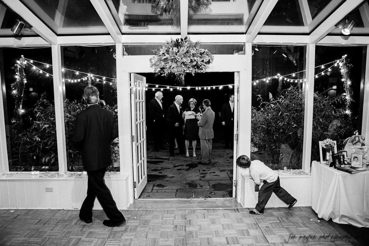 Black and white photo of a wedding reception. Guests gather outside a glass door adorned with lights and flowers. A child playfully peeks around the door, and a man walks inside the venue. Tables are set with decorations in the foreground.