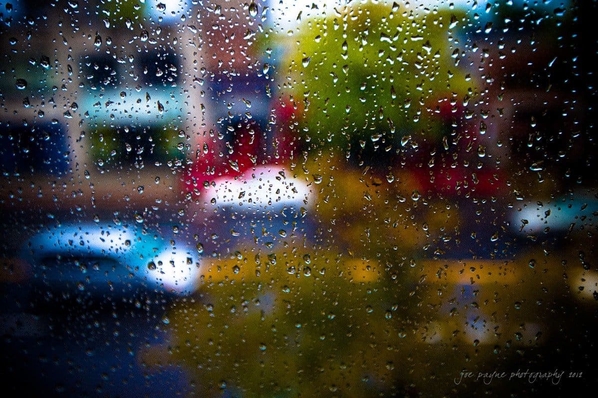Raindrops on a window with a blurred view of buildings, trees, and vehicles outside. The droplets create a textured effect on the glass, softly distorting the colorful street scene beyond.