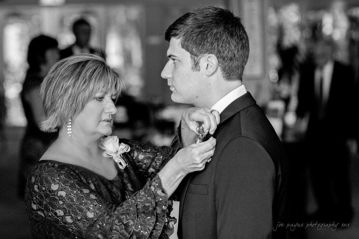 A woman is adjusting a boutonniere on a mans suit jacket. Both are dressed formally. The background is blurred, creating a focus on their interaction. The image is in black and white.