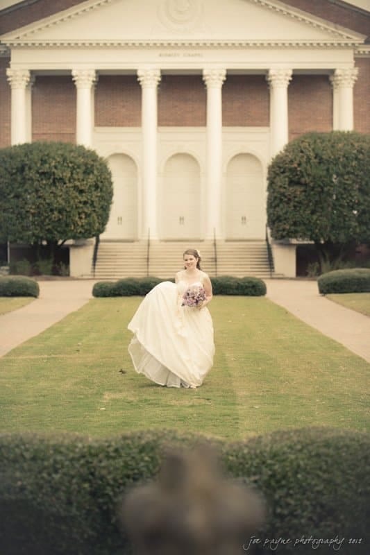 A bride in a white gown holds a bouquet while walking on a manicured lawn. Behind her is a brick building with large white columns. The scene is serene and elegant.