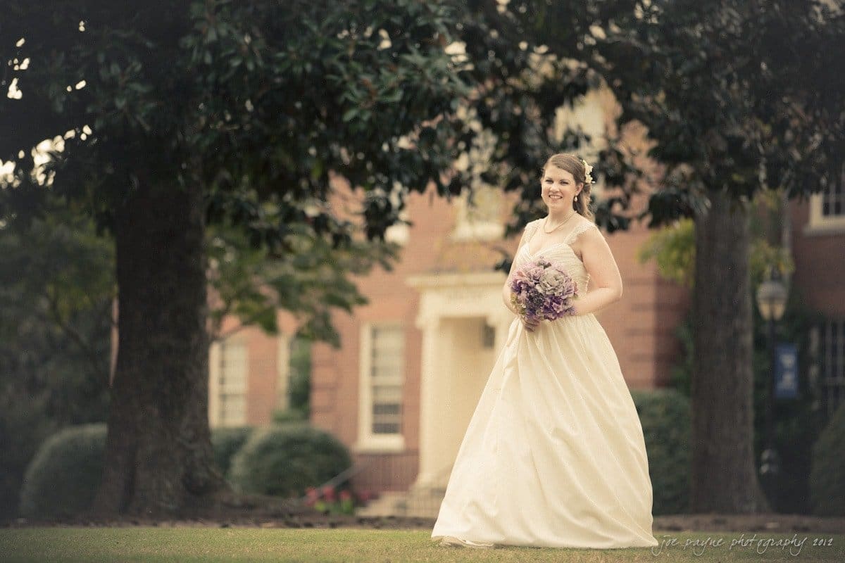 A bride in a flowing white gown holds a purple bouquet while walking across a lawn. She smiles as she moves past large trees, with a brick building in the background. The scene has a soft, romantic feel.