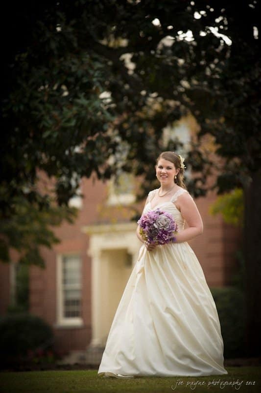 A bride in a white gown holds a bouquet of purple flowers, standing on a lawn with a blurred brick building in the background. She smiles under a large tree, with soft lighting casting a gentle glow.