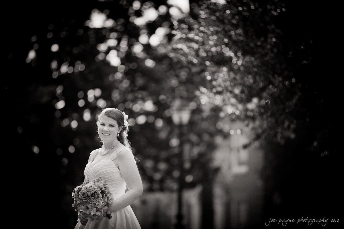 Bride standing outdoors in a glowing light, holding a bouquet and wearing a strapless gown. The background is softly blurred with trees and a lamppost visible. The image has a black and white, ethereal quality.