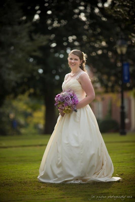 A bride in a white wedding dress stands on a grassy area, holding a bouquet of purple flowers. She smiles at the camera with a backdrop of trees and a lamp post. The lighting suggests a sunny day.