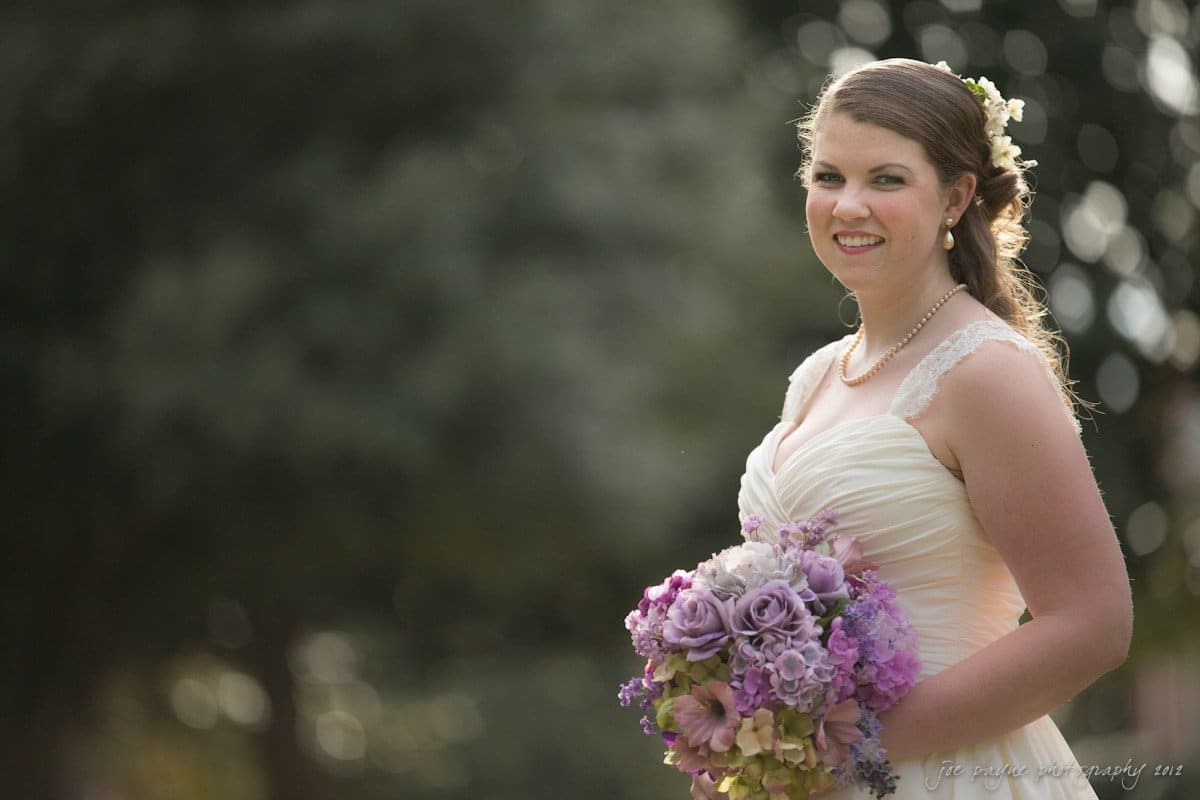 A bride in a white dress and floral hairpiece stands outdoors, smiling and holding a bouquet of purple and pink flowers. The background is softly blurred, highlighting the brides joyful expression and elegant attire.