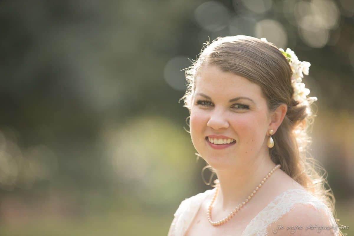 A woman in bridal attire smiles gently in an outdoor setting. She has a floral hair accessory and is wearing pearl earrings and a necklace. The background is softly blurred with warm lighting.