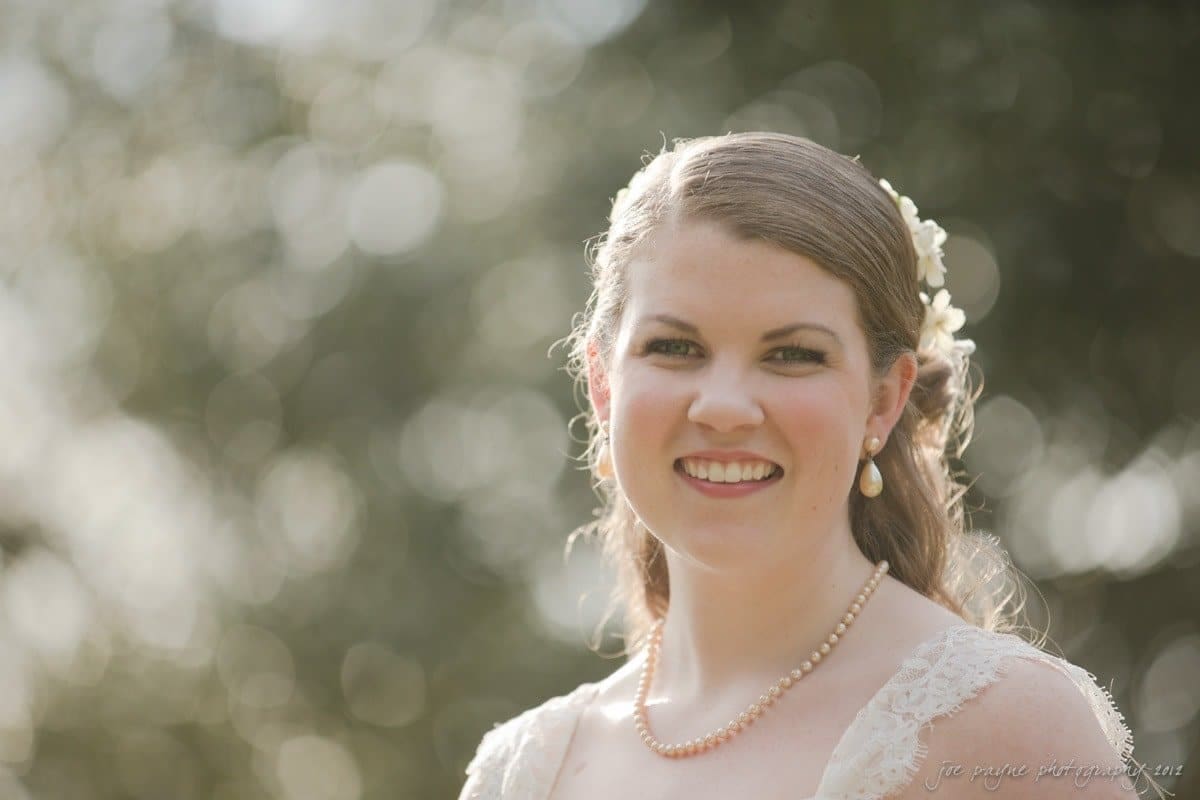 A woman with long hair adorned with flowers smiles warmly. She is wearing pearl earrings and a necklace, with a lace dress. The background is softly blurred, creating a gentle, dreamy atmosphere.